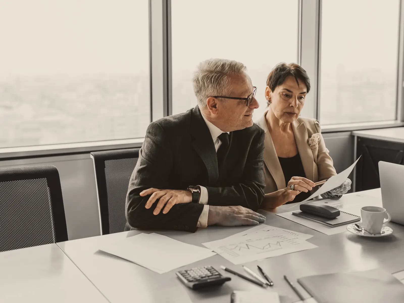 Two people check a printed page at a meeting table by a wide window; the people in colour, the room in black and white.