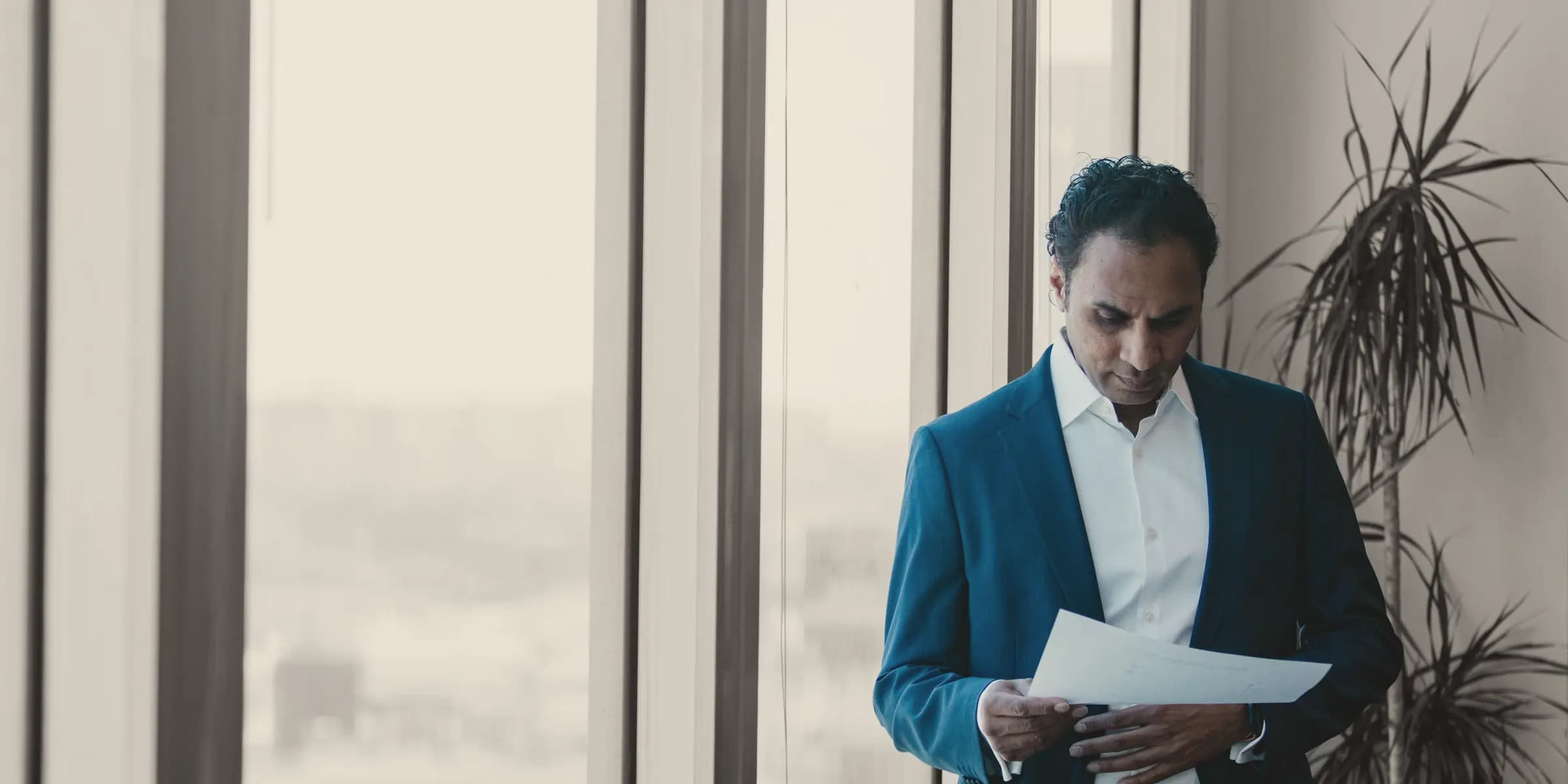 Someone in a blue suit reads a sheet by a tall window; the person in colour, the office in black and white.