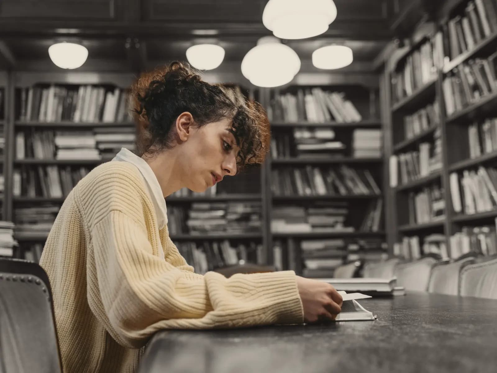 Someone reads at a long table in a library lined with books; the person in colour, the room in black and white.